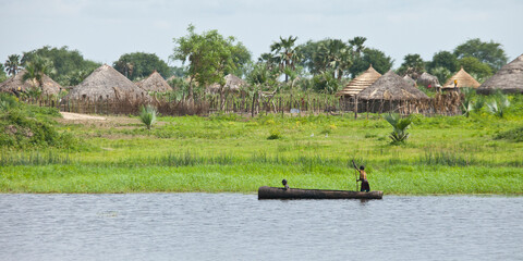 A dugout canoe goes past a village along the Nile River in South Sudan. - obrazy, fototapety, plakaty
