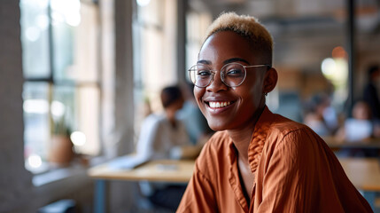 Joyful young woman with glasses is smiling broadly at the camera with a blurred office background