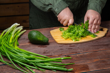 A woman's hand cuts greens in half with a knife on a wooden board on a table with vegetables