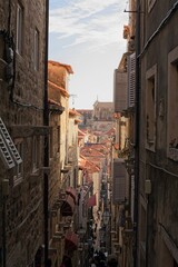 A bustling narrow street winds through the historic limestone buildings of Dubrovnik, Croatia, lined with red roofs, lanterns.