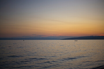 Dusk sets over the Adriatic Sea in Makarska, Croatia, a lone boat floats warm, orange-hued sky.
