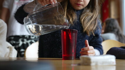 Child pouring water into red plastic cup with jar standing at restaurant, small girl serving refreshing tap water at diner