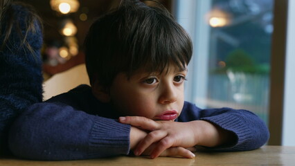 Pensive child feeling boredom at restaurant, leaning on table. Thoughtful little boy daydreaming, kid lost in thought