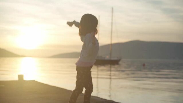 A girl on a pier by the sea throws stones into the water against the backdrop of sunset