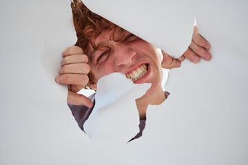 studio art portrait of a red-haired young man, tearing paper, peeking through a crack
