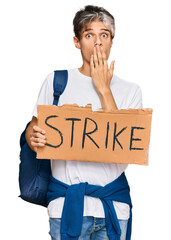 Young hispanic man holding strike banner cardboard covering mouth with hand, shocked and afraid for...