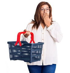 Middle age latin woman holding supermarket shopping basket covering mouth with hand, shocked and afraid for mistake. surprised expression