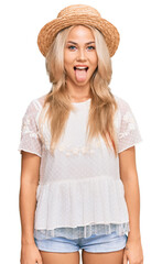 Young blonde girl wearing summer hat sticking tongue out happy with funny expression. emotion concept.