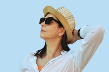 A brunette woman in a white shirt, sunglasses and a straw summer hat looks into the distance. Portrait of a woman traveler.