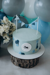 White flowers. Blue cake on a wooden stand  with decorative flags. The first cake. Cake on a white background. Blue, gray  and white balloons. Birthday. First birthday. Holiday