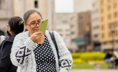 Fototapeta premium Latina adult woman, with a happy expression, in the city using her smartphone, sending audio messages and typing. Copyspace......