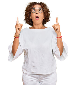 Beautiful middle age mature woman wearing casual clothes and glasses amazed and surprised looking up and pointing with fingers and raised arms.