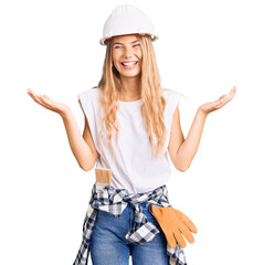 Beautiful caucasian woman with blonde hair wearing hardhat and painter clothes celebrating mad and...