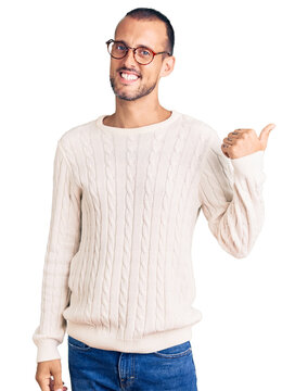 Young handsome man wearing casual clothes and glasses smiling with happy face looking and pointing to the side with thumb up.