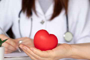 Patient holding red heart in hand with female doctor writing in the background. Organ donation, cardiology National Day, love, compassion, healthcare concept. - obrazy, fototapety, plakaty