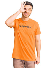 Handsome young man with bear wearing tshirt with happiness word message doing ok gesture with hand smiling, eye looking through fingers with happy face.