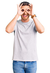 Handsome young man with bear wearing casual tshirt doing ok gesture like binoculars sticking tongue out, eyes looking through fingers. crazy expression.