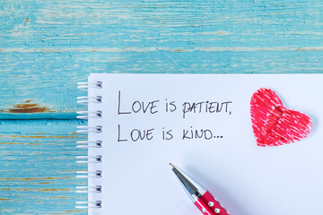 Love is patient and kind, handwritten verse in notebook with pen and red paper heart on wood....