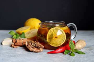 Teapot with hot tea, ginger, turmeric root, cinnamon sticks, lemon and mint leaves on grey...