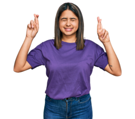 Young hispanic girl wearing casual purple t shirt gesturing finger crossed smiling with hope and eyes closed. luck and superstitious concept.