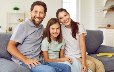 Portrait of smiling young happy family of three with daughter sitting on the sofa at home and looking cheerful at camera. Child girl sitting with parents. Love, care, family time and leisure concept.