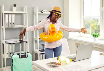 Funny happy smiling african american woman standing at the desk on workplace at office in a beach rubber ring with suitcase and booking tickets for summer vacation online via laptop. - obrazy, fototapety, plakaty