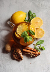 Teapot with hot tea, ginger, turmeric root, cinnamon sticks, lemon and mint leaves on grey background with copy space. Anti inflammatory winter drink recipe. 