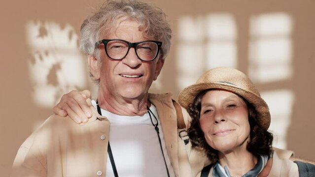Lovely frontal view of older Caucasian couple standing together and hugging each other. People looking directly at camera while sunlight brightens their faces. Stylish, elegant look.