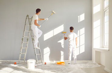 Back view of a team of professional painters in white clothes painting a grey wall in empty room...