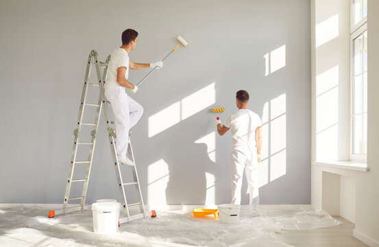 Back view of a team of professional painters in white clothes painting a grey wall in empty room with paint roller. Building contractors doing repair renovation in client house. Repair concept.