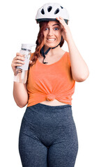 Young beautiful woman wearing bike helmet and holding water bottle stressed and frustrated with hand on head, surprised and angry face
