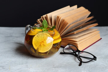 Glass teapot with ginger and lemon tea, open book and reading glasses on a table. Winter day still...