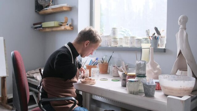 a man with hand pathology paints pottery with glaze in the workshop.