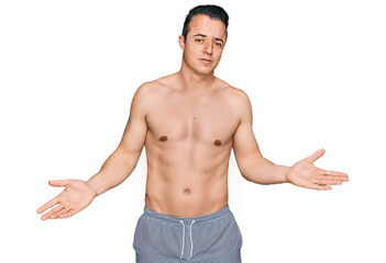 Handsome young man wearing swimwear shirtless clueless and confused with open arms, no idea concept.