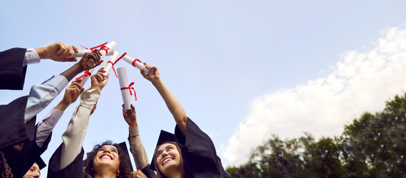 Bottom view portrait of a group of smiling happy graduates students raising their diplomas up to the sky standing in a circle in graduate gown. Education and graduation concept. Banner.