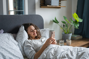 Young smiling woman lying in bed in the morning and watching news and social media on her smartphone