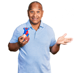 Hispanic middle age man holding heart organ celebrating achievement with happy smile and winner expression with raised hand