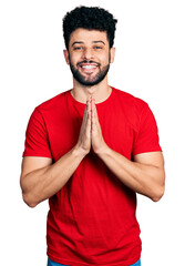 Young arab man with beard wearing casual red t shirt praying with hands together asking for...