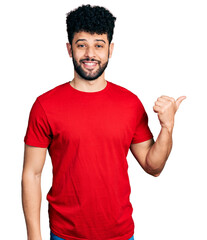 Young arab man with beard wearing casual red t shirt smiling with happy face looking and pointing...