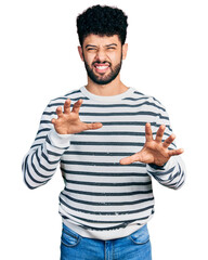 Young arab man with beard wearing casual striped sweater smiling funny doing claw gesture as cat,...
