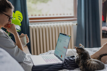Young woman with cute gray kitten working on a project using laptop in bed at home