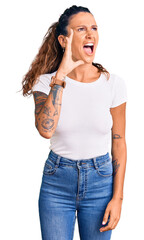 Young hispanic woman with tattoo wearing casual white tshirt shouting and screaming loud to side with hand on mouth. communication concept.
