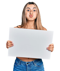 Hispanic young woman holding blank empty banner puffing cheeks with funny face. mouth inflated with air, catching air.
