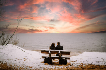 couple sitting on a bench by the sea, with a snowy beach in the foreground and a colorful sunset sky.