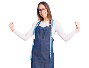 Beautiful brunette young woman dressmaker designer wearing atelier apron very happy and excited doing winner gesture with arms raised, smiling and screaming for success. celebration concept.