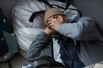 High angle portrait of distressed young man sleeping on floor at refugee shelter and covering face with hands