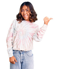 Young beautiful mixed race woman wearing casual tie dye sweatshirt smiling with happy face looking and pointing to the side with thumb up.