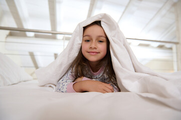 Close-up portrait of a little child girl 6 years old lying on the bed under white blanket while waking up in the morning