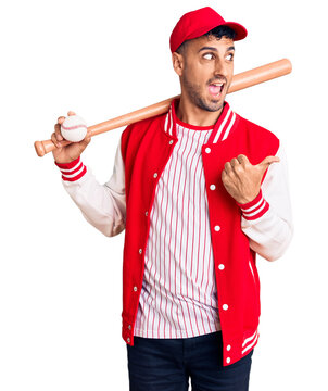 Young hispanic man playing baseball holding bat and ball pointing thumb up to the side smiling happy with open mouth
