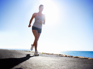 Woman, fitness and running on beach for workout, exercise or outdoor training on a sunny day. Female person, athlete or runner in race, sprint or sports marathon on asphalt, road by the ocean coast